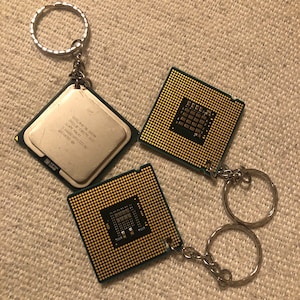 May include: Two gold-colored computer chip keychains with silver key rings. The chips have a green circuit board design and the text "Intel Core i7 CPU" is visible on one of the chips.