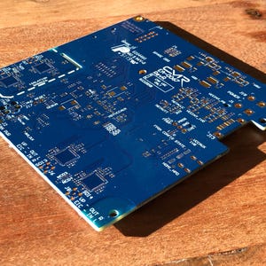 Puede incluir: Una placa de circuito impreso azul con componentes y texto de color dorado. El texto incluye "CVR" y "Reve" y una serie de números. La placa tiene una forma rectangular con esquinas redondeadas.