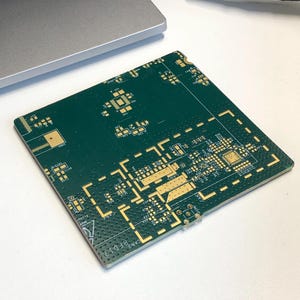 Może przedstawiać: Zielona płytka drukowana z pozłacanymi ścieżkami i komponentami. Płytka ma kształt prostokąta i wymiary około 10 cm na 10 cm. Płytka ma etykietę z napisem "CANDY PBD17".