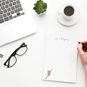 Può includere: Un blocco note bianco con la parola "notizen" scritta su di esso. Una mano sta scrivendo sul blocco note con una penna nera. C'è una calcolatrice, un paio di occhiali, un laptop, una pianta in vaso e una tazza di caffè su una superficie bianca.