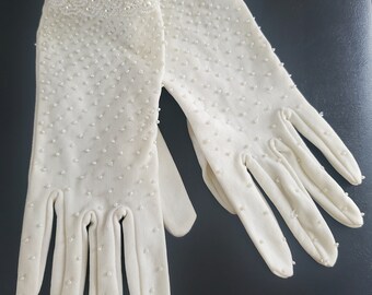May include: A pair of white gloves with pearl embellishments. The gloves have a beaded trim around the wrist and are covered in small pearls.