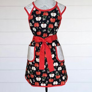 May include: A black and red apron with a white polka dot ribbon and apple print. The apron has two large pockets on the front.
