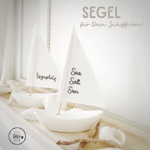 Könnte beinhalten: Zwei weiße Miniatur-Segelschiffe mit Stoffsegeln. Das Segel links sagt "liegeplatz" und das Segel rechts sagt "Sea Salt Sun".