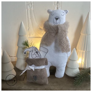 May include: A white polar bear plush toy wearing a brown faux fur scarf, standing next to a brown burlap sack with a white trim and a white and gray embroidered design. The sack is filled with a white and gray plush toy with a red hat. The background features white decorative trees.