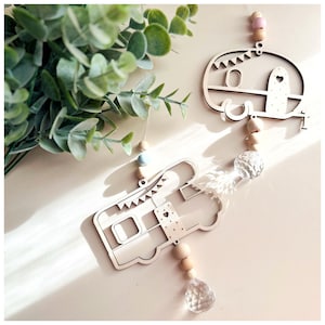 May include: Two wooden camper van ornaments hanging from a string with wooden beads and crystal pendants. The camper vans are painted in a light wood color and have a white heart on the side.