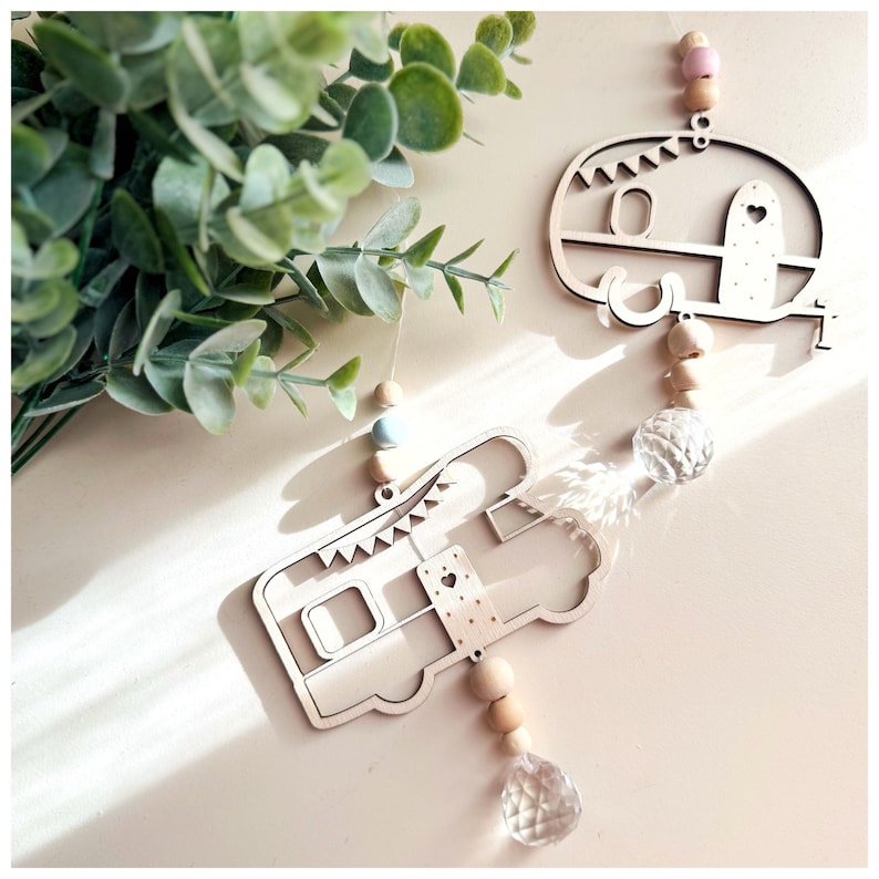 May include: Two wooden camper van ornaments hanging from a string with wooden beads and crystal pendants. The camper vans are painted in a light wood color and have a white heart on the side.