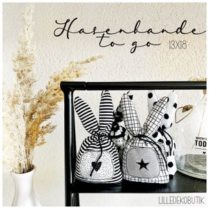 Puede incluir: Tres bolsas de cordón con forma de conejo en blanco y negro con diferentes patrones. Las bolsas están colgadas en un estante de metal negro. El texto "Hasenbande to go 13x18" está escrito en una pared blanca detrás de las bolsas.