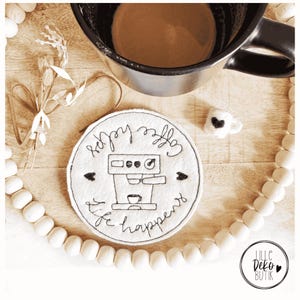 Può includere: Un sottobicchiere in feltro bianco con la scritta "Coffee helps life happens" e l'illustrazione di una macchina da caffè. Sono presenti anche una tazza da caffè nera, una piccola tazza in feltro e perline decorative. Il sottobicchiere misura circa 10 cm di diametro.