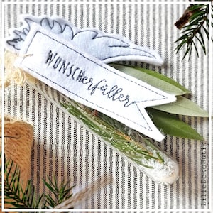 Puede incluir: Un pequeño tubo de vidrio lleno de agujas de pino y purpurina blanca, con una etiqueta de fieltro blanco que dice "Wunsch-erfüller" y un ala de fieltro azul adjunta.