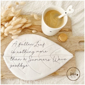 Könnte beinhalten: Ein weißer, blattförmiger Untersetzer mit dem gestickten Text "A fallen leaf is nothing more than a Summers Wave goodbye". Der Untersetzer liegt auf einer Holzoberfläche mit einer Tasse Kaffee, einem Löffel und einem Macaron.