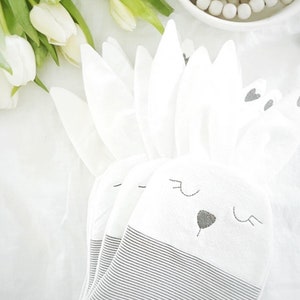 Könnte beinhalten: Weiße, hasenförmige Servietten mit schwarzen und grauen Akzenten. Die Servietten haben ein gestreiftes Muster am unteren Rand und gestickte Gesichtsmerkmale. Weiße Tulpen sind im Hintergrund.