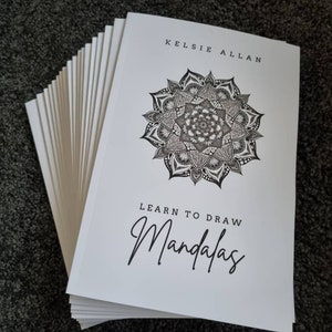 May include: A stack of white paperbacks with a black and white mandala design on the cover. The title of the book is "Learn to Draw Mandalas" and the author is "Kelsei Allan".