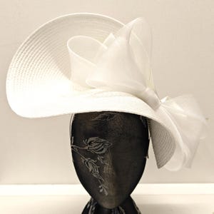 Peut inclure: Chapeau blanc à larges bords orné d'un grand nœud décoratif en tissu transparent. Le chapeau est en tissu tissé et est présenté sur une tête de mannequin noire avec un motif floral.