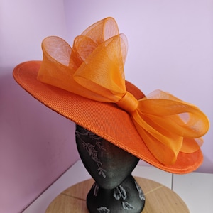 Könnte beinhalten: Ein leuchtend orangefarbener Hut mit breiter Krempe und einer großen, mehrlagigen Schleife. Die Schleife besteht aus einem transparenten, strukturierten Stoff, der einen zarten Kontrast zur einfarbigen Farbe des Hutes bildet. Der Hut wird auf einem schwarzen Mannequinkopf präsentiert.