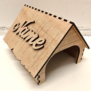 May include: A wooden house-shaped sign with a cutout for a name. The sign is made of light brown wood and has a black border. The word "Name" is cut out of the wood and is in a cursive font.