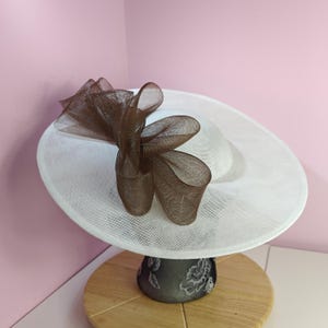 May include: A white hat with a brown mesh bow on top. The hat is on a display stand.