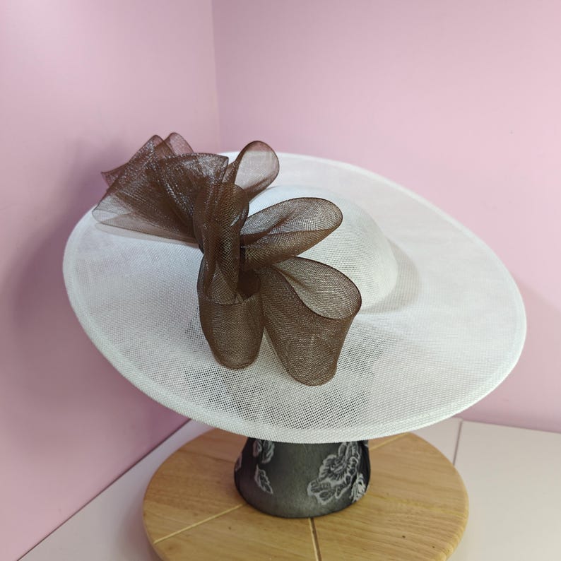 May include: A white hat with a brown mesh bow on top. The hat is on a display stand.