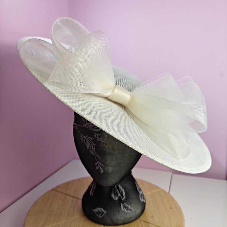 May include: A white hat with a large white bow. The hat is made of a stiff material and has a wide brim. The bow is made of a soft, flowing material.