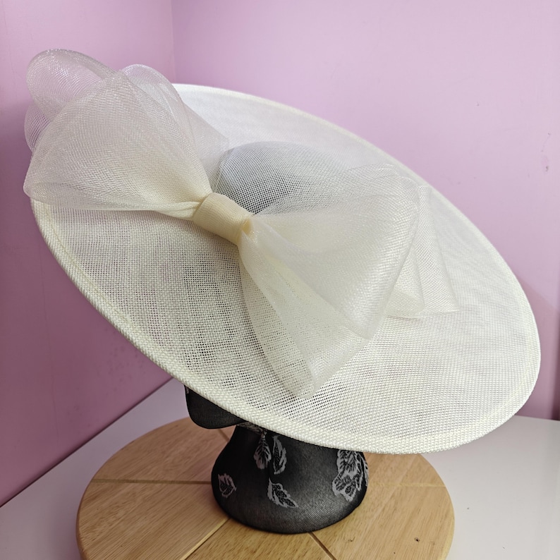 May include: A large, white, wide-brimmed hat with a large white bow. The hat is made of a fine, woven material and is sitting on a black mannequin head.