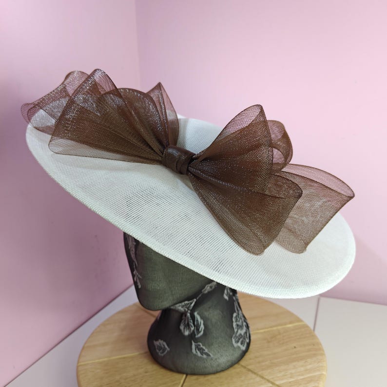 May include: A white hat with a large brown bow made of tulle. The hat is sitting on a mannequin head.