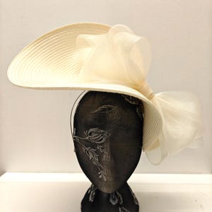 Könnte beinhalten: Ein großer, cremefarbener Hut mit breiter Krempe und einer großen, cremefarbenen Schleife an der Seite.