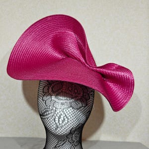 Può includere: Un cappello fucsia vibrante con una tesa ampia e asimmetrica. Il cappello è realizzato in un materiale tessuto, con una superficie strutturata. Il cappello è esposto su una testa di manichino nera con una copertura in pizzo. Un accessorio elegante.