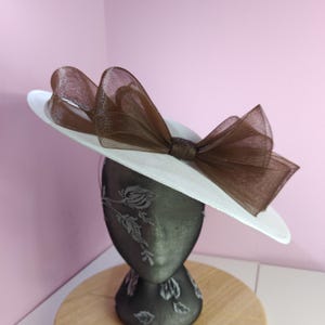 May include: A white hat with a large brown bow. The hat is on a black mannequin head.