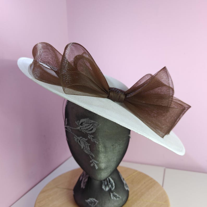May include: A white hat with a large brown bow. The hat is on a black mannequin head.