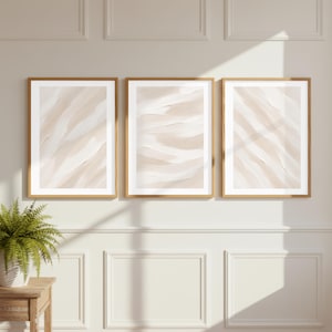 May include: A set of three vertical wall art prints in light wood frames, each featuring abstract, textured brushstrokes in shades of cream, beige, and white, displayed side-by-side on a wall with paneling and a green potted plant nearby.