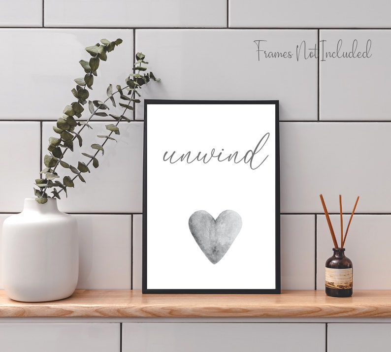May include: A black framed print with the word "unwind" in cursive script and a gray watercolor heart. The print is on a white background and is displayed on a shelf with a vase of greenery and a diffuser.