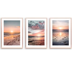 May include: A set of three vertical wall art prints in thin rose gold frames. Each print features a different beach sunset scene with soft pink, orange, and blue hues, showing ocean waves, sandy shores, and reflective water surfaces.