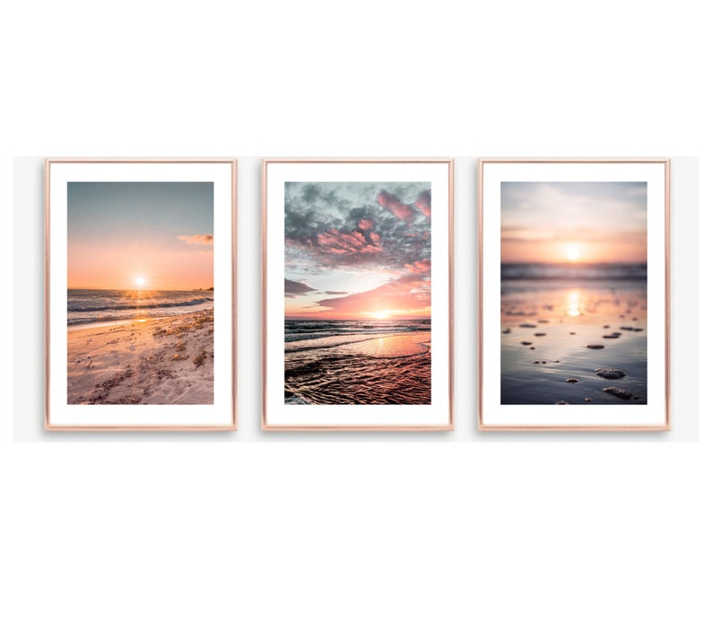 May include: A set of three vertical wall art prints in thin rose gold frames. Each print features a different beach sunset scene with soft pink, orange, and blue hues, showing ocean waves, sandy shores, and reflective water surfaces.