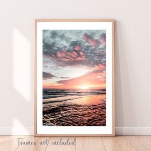 May include: A vertical wall art print featuring a sunset over the ocean with pink and orange hues in the sky and clouds reflecting on the wet sand. The print is displayed in a light wood frame with a white mat. The text "Frames not included" is visible.