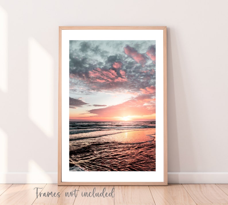 May include: A vertical wall art print featuring a sunset over the ocean with pink and orange hues in the sky and clouds reflecting on the wet sand. The print is displayed in a light wood frame with a white mat. The text "Frames not included" is visible.