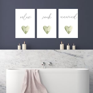 May include: Three white framed prints with green watercolor hearts and the words "relax", "soak", and "unwind" in black calligraphy. The prints are hanging on a dark blue wall above a white bathtub.