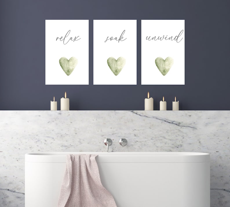 May include: Three white framed prints with green watercolor hearts and the words "relax", "soak", and "unwind" in black calligraphy. The prints are hanging on a dark blue wall above a white bathtub.