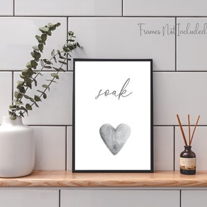 May include: A black framed print with the word "soak" in cursive script and a gray watercolor heart. The print is on a white background and is sitting on a wooden shelf.