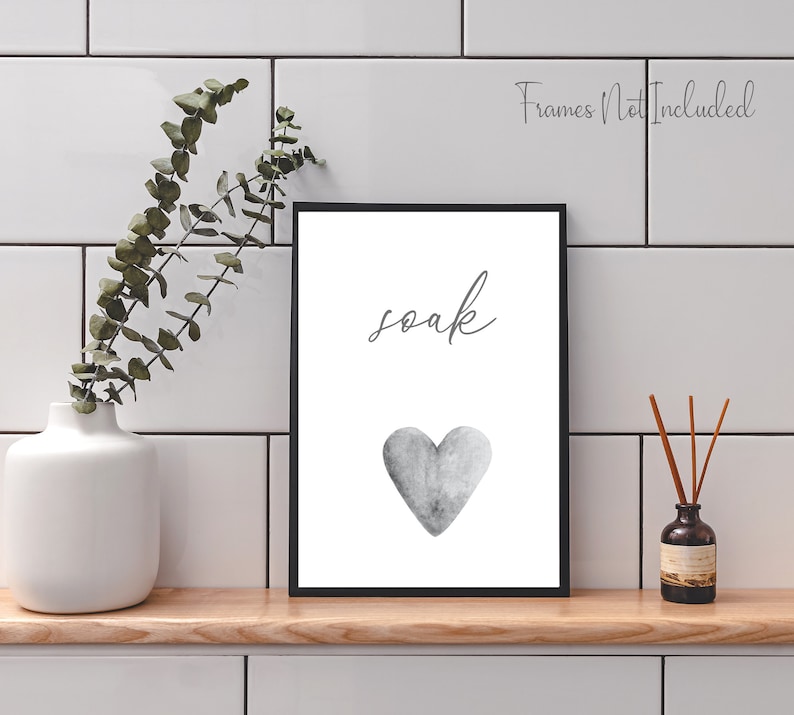 May include: A black framed print with the word "soak" in cursive script and a gray watercolor heart. The print is on a white background and is sitting on a wooden shelf.