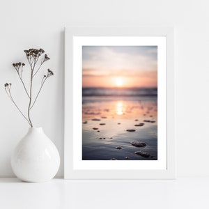 May include: A white framed wall art print featuring a sunset over a beach with wet sand and small bubbles, displayed next to a white ceramic vase containing a single dried plant stem with small dark brown seed pods on thin branches.