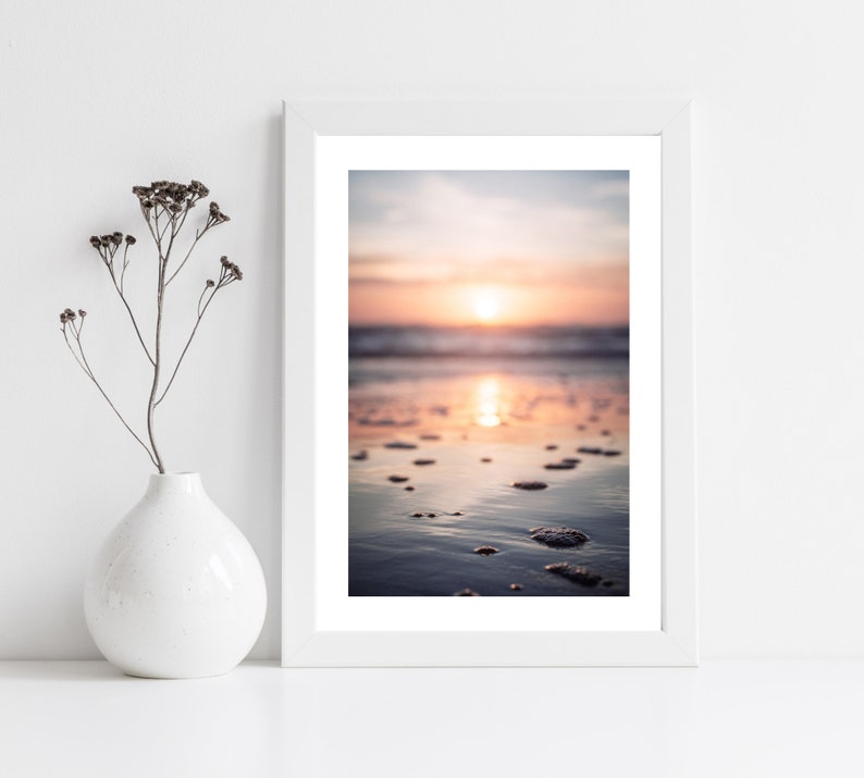May include: A white framed wall art print featuring a sunset over a beach with wet sand and small bubbles, displayed next to a white ceramic vase containing a single dried plant stem with small dark brown seed pods on thin branches.