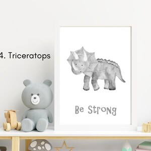 Puede incluir: Una ilustraci&oacute;n de acuarela en blanco y negro de un dinosaurio Triceratops con el texto "Be Strong" impreso debajo. La ilustraci&oacute;n est&aacute; enmarcada en un marco blanco y se muestra en un estante con un oso de peluche gris y otros art&iacute;culos decorativos.