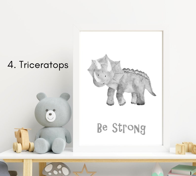 Puede incluir: Una ilustraci&oacute;n de acuarela en blanco y negro de un dinosaurio Triceratops con el texto "Be Strong" impreso debajo. La ilustraci&oacute;n est&aacute; enmarcada en un marco blanco y se muestra en un estante con un oso de peluche gris y otros art&iacute;culos decorativos.