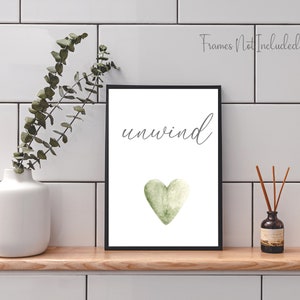 May include: A white printable art print with the word "unwind" in black cursive script and a green watercolor heart. The print is framed in a black frame and is displayed on a white tiled wall.