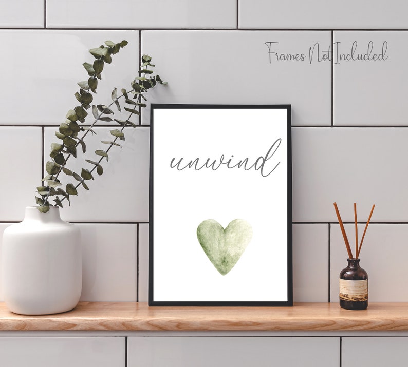 May include: A white printable art print with the word "unwind" in black cursive script and a green watercolor heart. The print is framed in a black frame and is displayed on a white tiled wall.