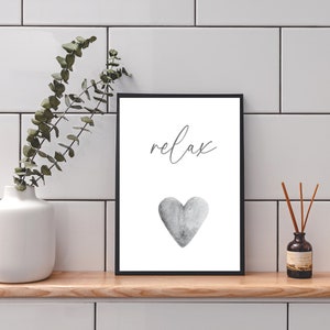 May include: A black framed print with the word "relax" in cursive script and a gray watercolor heart. The print is on a white background and is sitting on a wooden shelf.