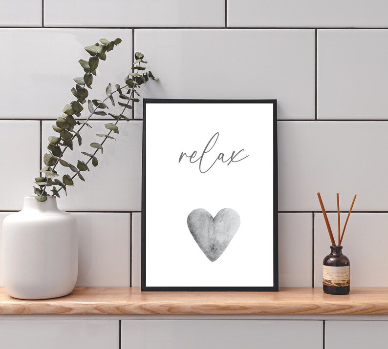 May include: A black framed print with the word "relax" in cursive script and a gray watercolor heart. The print is on a white background and is sitting on a wooden shelf.
