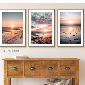 May include: Three vertical wall art prints featuring beach sunset scenes with orange, pink, and blue hues. The left print shows a sandy shore, the center shows ocean waves under a cloudy sky, and the right shows a blurred shoreline with water reflections.