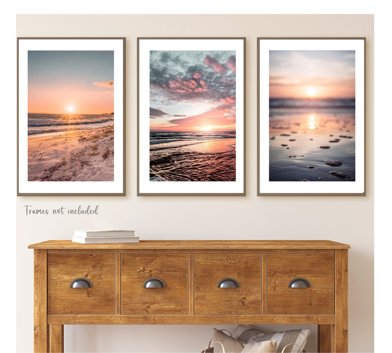 May include: Three vertical wall art prints featuring beach sunset scenes with orange, pink, and blue hues. The left print shows a sandy shore, the center shows ocean waves under a cloudy sky, and the right shows a blurred shoreline with water reflections.
