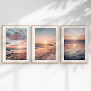 May include: Three vertical framed wall art prints featuring beach sunset scenes. The left print shows pink clouds over the ocean, the center print displays a sunlit sandy shore, and the right print captures a blurred shoreline with wet sand reflections.