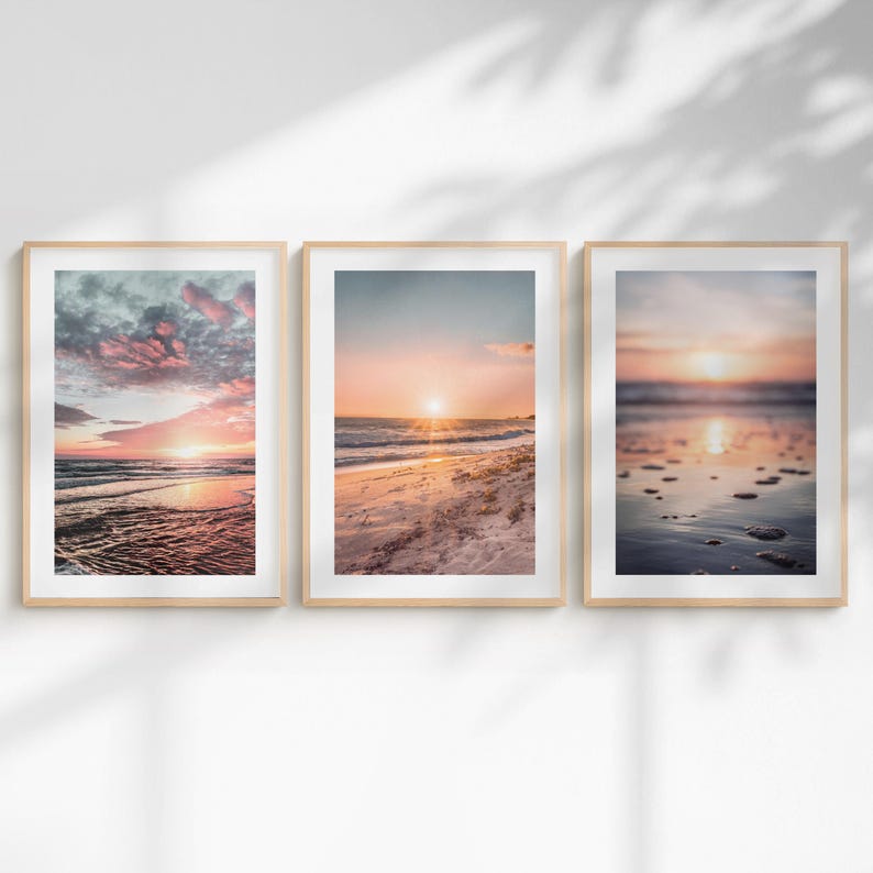 May include: Three vertical framed wall art prints featuring beach sunset scenes. The left print shows pink clouds over the ocean, the center print displays a sunlit sandy shore, and the right print captures a blurred shoreline with wet sand reflections.